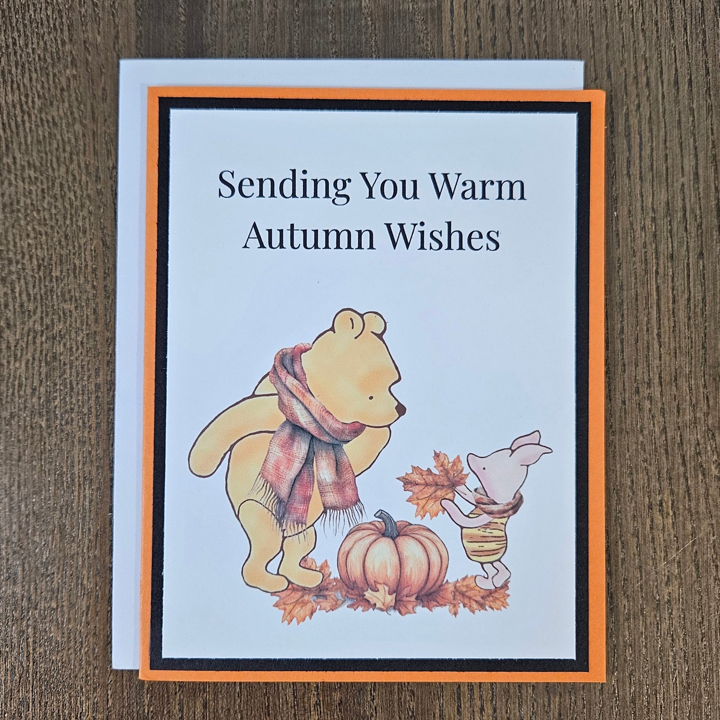 Front of a handmade A2 autumn card featuring classic Winnie the Pooh and Piglet wearing scarves next to a pumpkin framed by black and orange borders with text reading Sending You Warm Autumn Wishes.