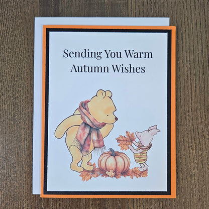 Front of a handmade A2 autumn card featuring classic Winnie the Pooh and Piglet wearing scarves next to a pumpkin framed by black and orange borders with text reading Sending You Warm Autumn Wishes.