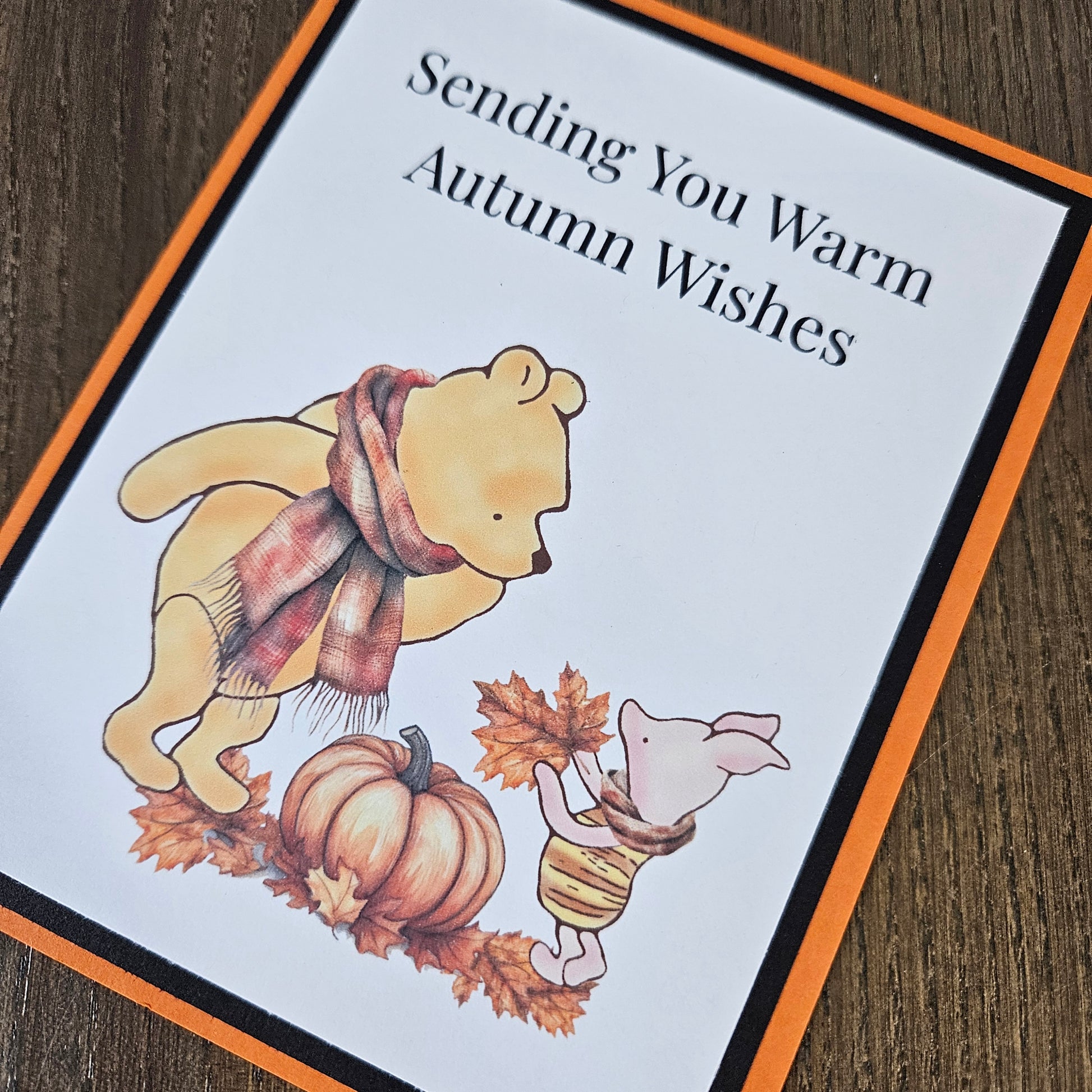 An autumn-themed greeting card featuring Winnie the Pooh and Piglet with a pumpkin and leaves, bordered in orange and black, placed on a wooden surface.