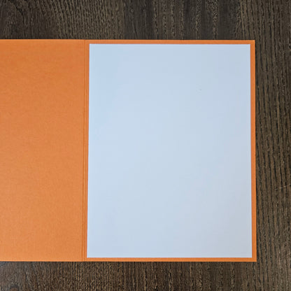 An open orange greeting card or notebook with a blank white page on the right, photographed from above on a dark wood surface.