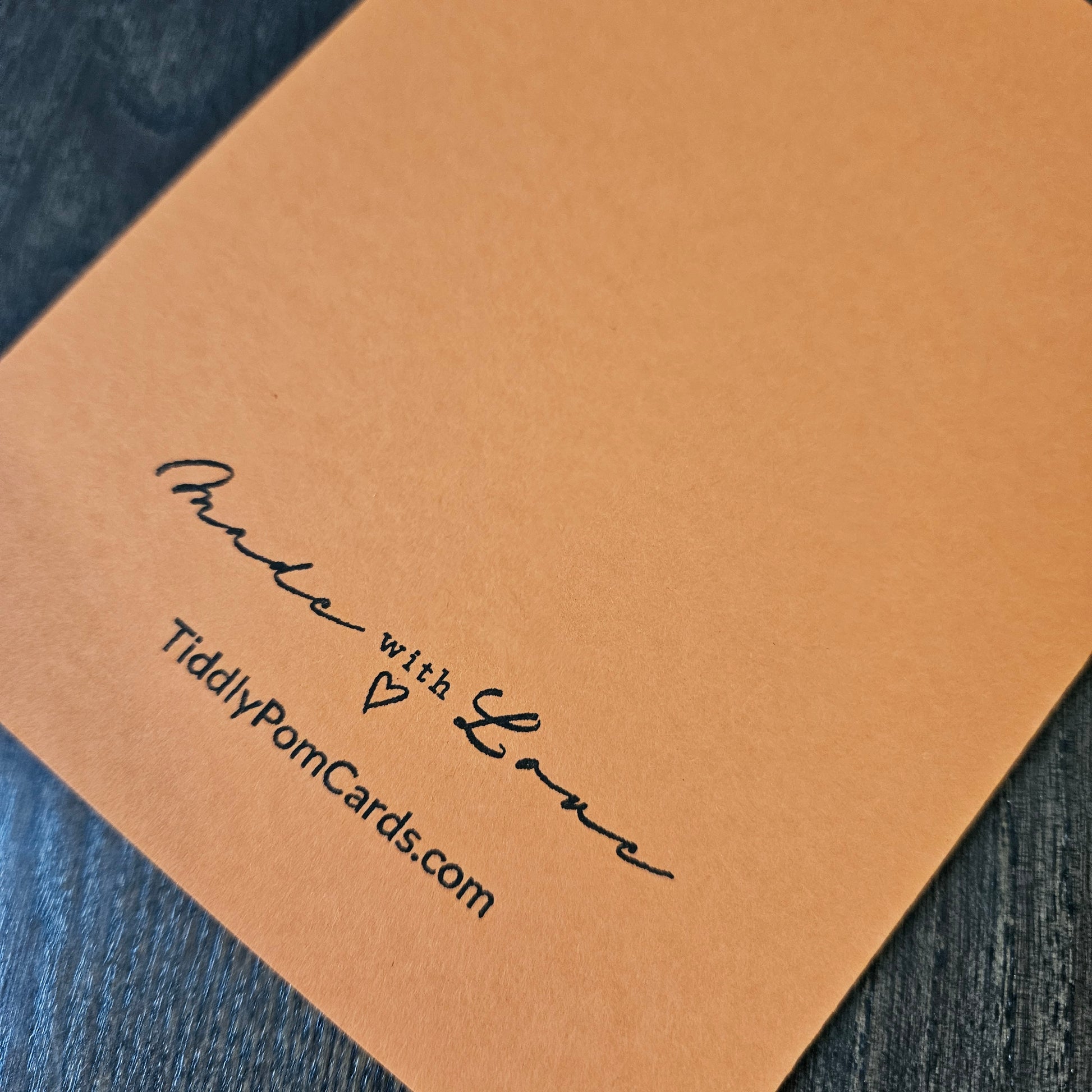A close-up of a square tan pom pom card with black printed text reading 'Made with Love' and 'TiddlyPomCards.com', placed on a dark textured surface.