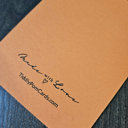 A close-up of a square tan pom pom card with black printed text reading 'Made with Love' and 'TiddlyPomCards.com', placed on a dark textured surface.