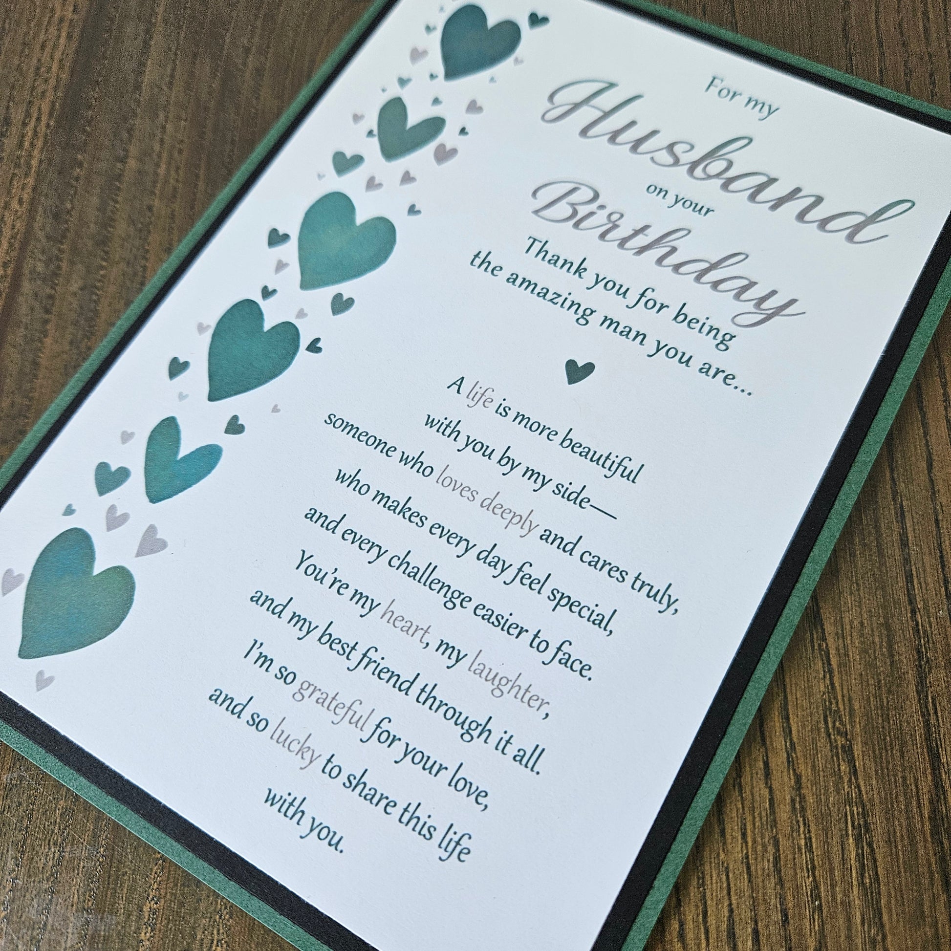 A square greeting card for a husband's birthday, featuring a white background with teal and gray hearts, elegant gray script text, and a dark green border, displayed on a wooden surface.