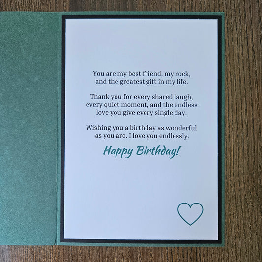 A folded greeting card shown open on a wooden surface, with a teal-green card backing and a white front panel printed with a birthday message and a small heart outline.