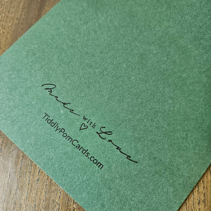 A close-up of a green textured card or envelope corner printed with the words 'Make with Love' and 'TiddlyPornCards.com' in black script, placed on a wooden surface.