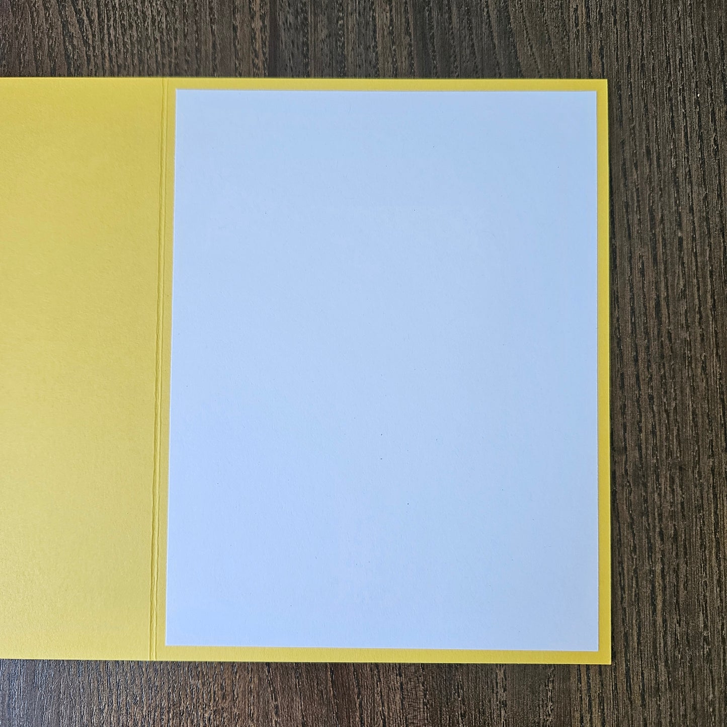 A blank greeting card with a yellow border and a light blue center panel, shown open on a dark wood background.