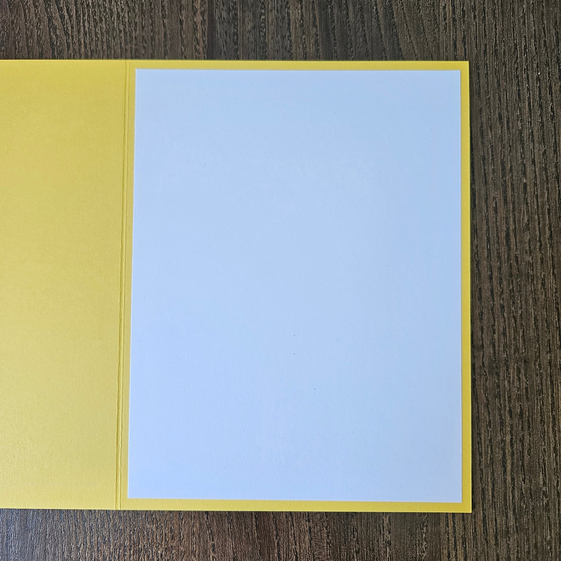 A blank greeting card with a yellow border and a light blue center panel, shown open on a dark wood background.