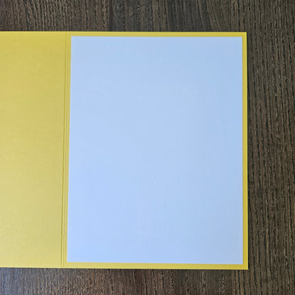 A blank greeting card with a yellow border and a light blue center panel, shown open on a dark wood background.