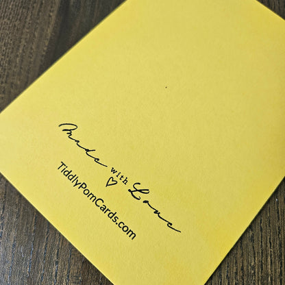 A close-up of a yellow greeting card or paper product placed on a dark wood surface, printed with the words 'Make with Love' and 'TidilyPomCards.com' in black script.