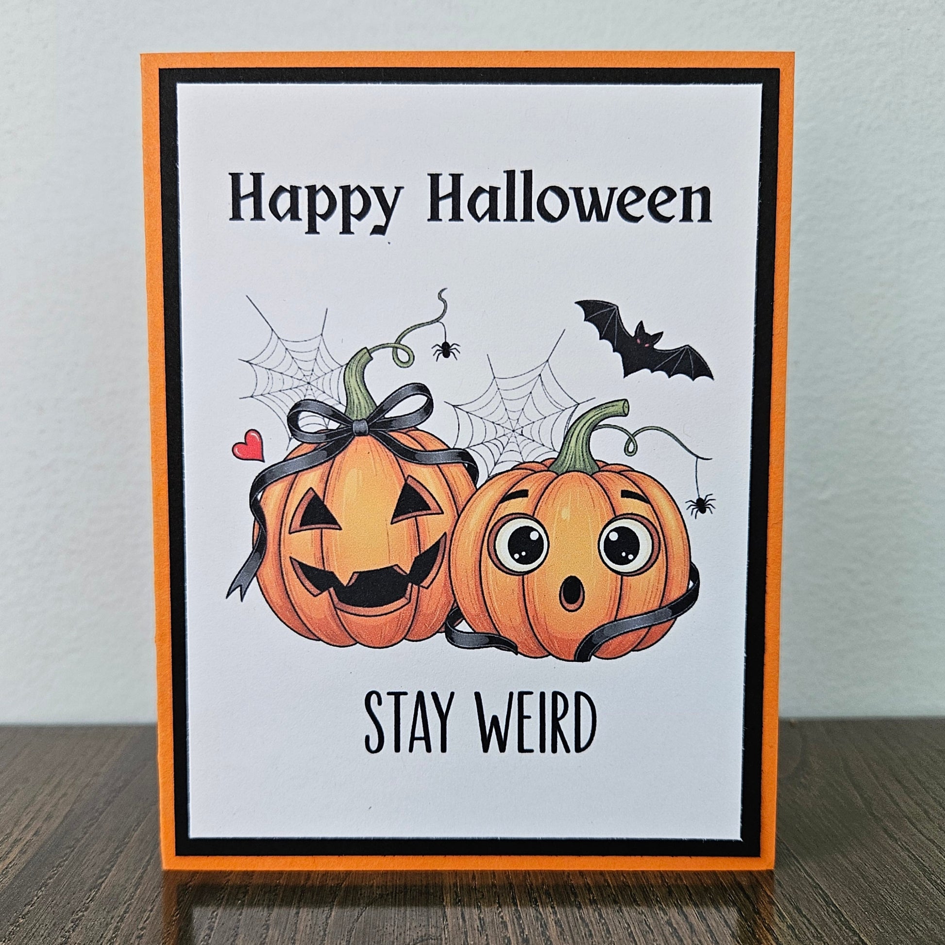 Front of a handmade A2 Halloween card featuring two illustrated pumpkins with ribbon bows, spider webs, and a bat framed by black and orange borders reading Happy Halloween STAY WEIRD.