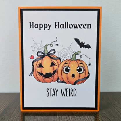 Front of a handmade A2 Halloween card featuring two illustrated pumpkins with ribbon bows, spider webs, and a bat framed by black and orange borders reading Happy Halloween STAY WEIRD.