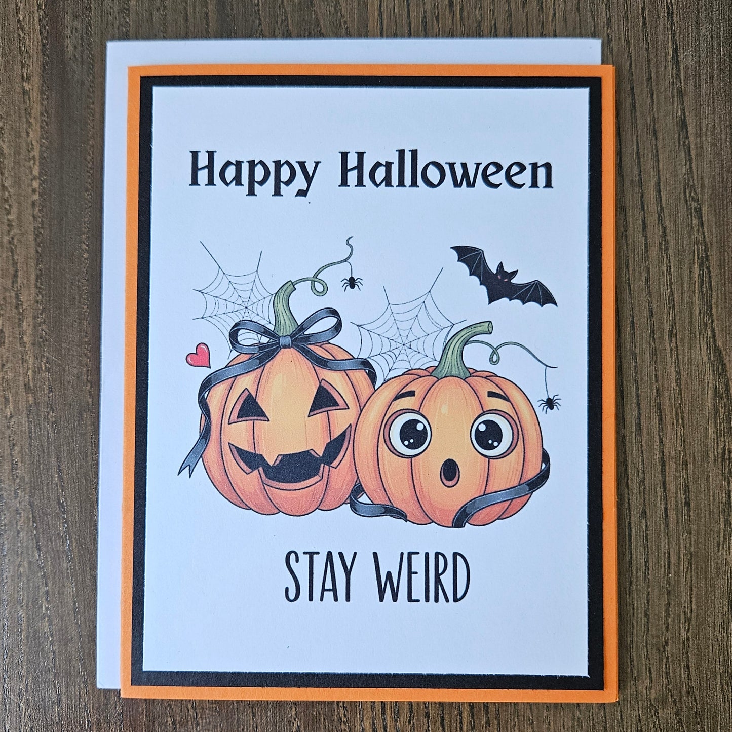 A Halloween greeting card with a white front, orange border, and black inner frame, placed on a brown wood surface. The card reads 'Happy Halloween' and 'STAY WEIRD' with cartoon pumpkins, spiderwebs, a bat, and small hearts.
