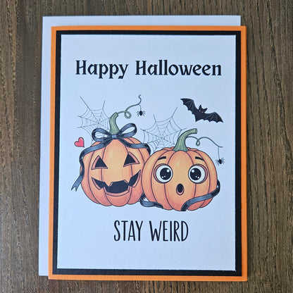 A Halloween greeting card with a white front, orange border, and black inner frame, placed on a brown wood surface. The card reads 'Happy Halloween' and 'STAY WEIRD' with cartoon pumpkins, spiderwebs, a bat, and small hearts.