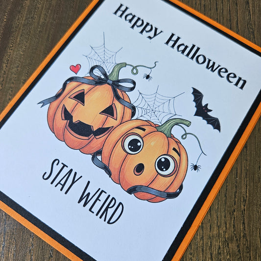 A Halloween greeting card with an orange border, featuring two cartoon jack-o'-lanterns, spiderwebs, a bat, and the text 'Happy Halloween' and 'STAY WEIRD' on a white background, photographed on a wooden surface.