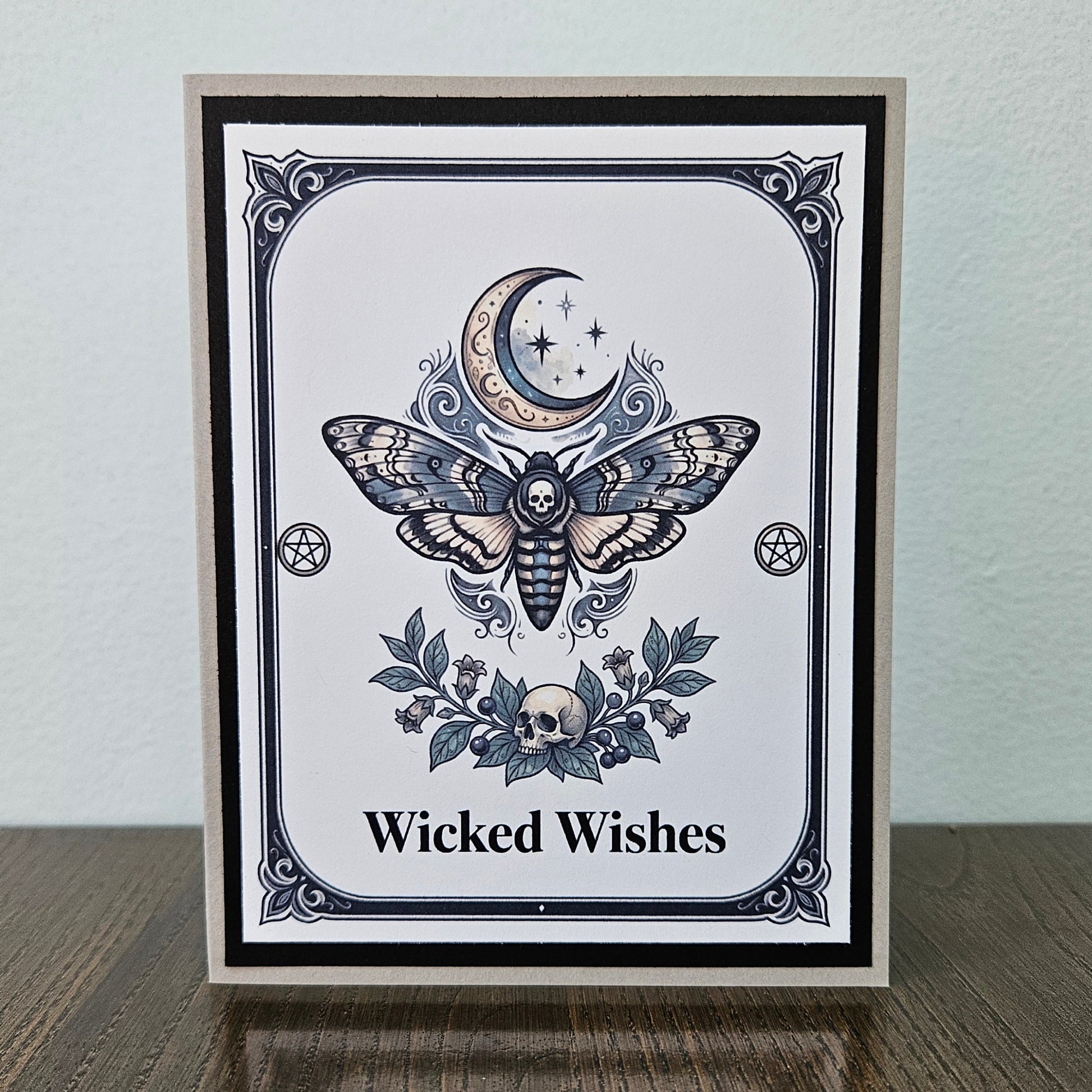 Front of a handmade A2 gothic card featuring a death head moth under a crescent moon with skulls and pentagrams framed in black and gray with text reading Wicked Wishes.