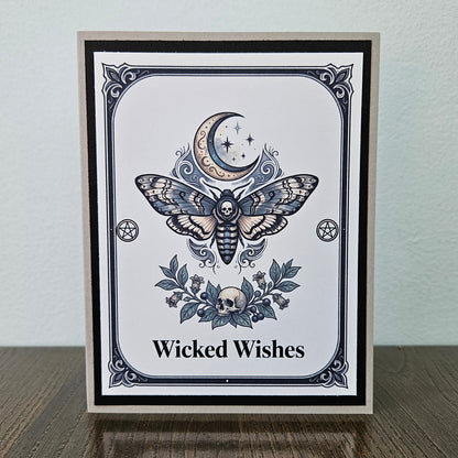 Front of a handmade A2 gothic card featuring a death head moth under a crescent moon with skulls and pentagrams framed in black and gray with text reading Wicked Wishes.