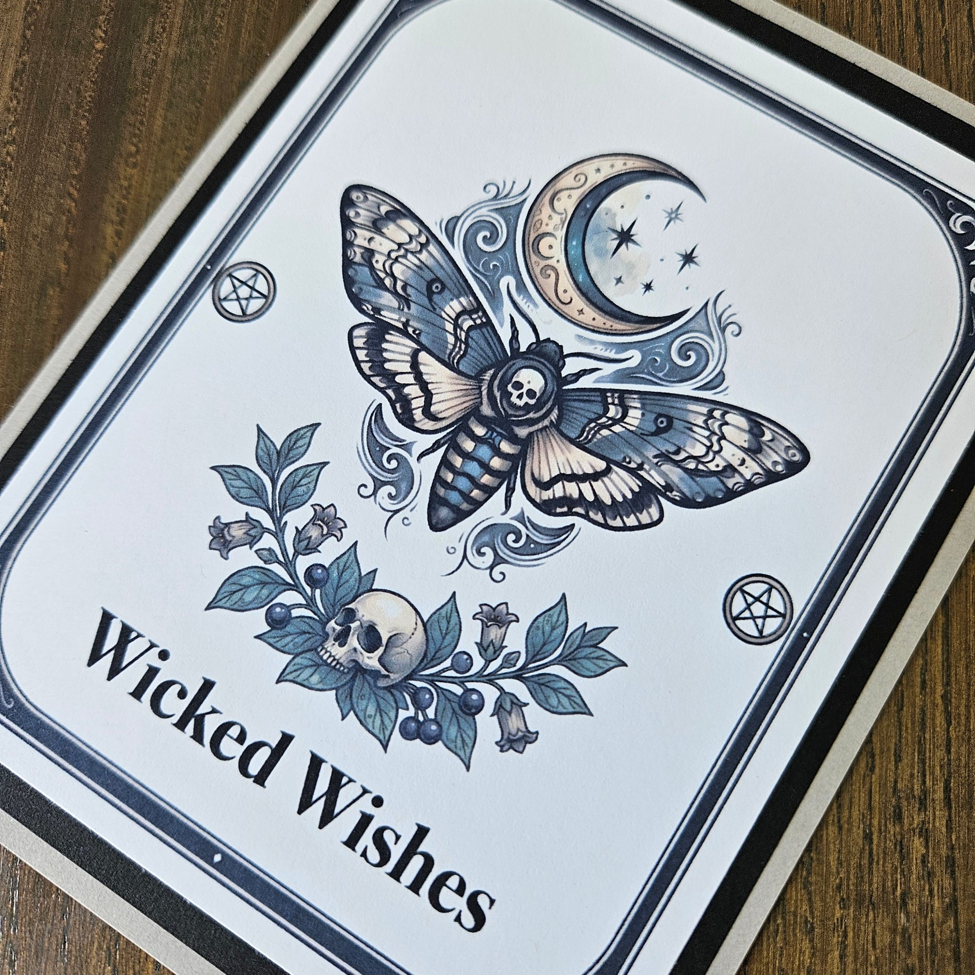 A square printed card or sign with a white background and black border, featuring a detailed illustration of a moth with a skull in its wings, a crescent moon with stars, floral elements, and the text 'Wicked Wishes.' The card is photographed on a wooden surface.