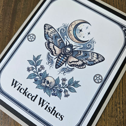 A square printed card or sign with a white background and black border, featuring a detailed illustration of a moth with a skull in its wings, a crescent moon with stars, floral elements, and the text 'Wicked Wishes.' The card is photographed on a wooden surface.