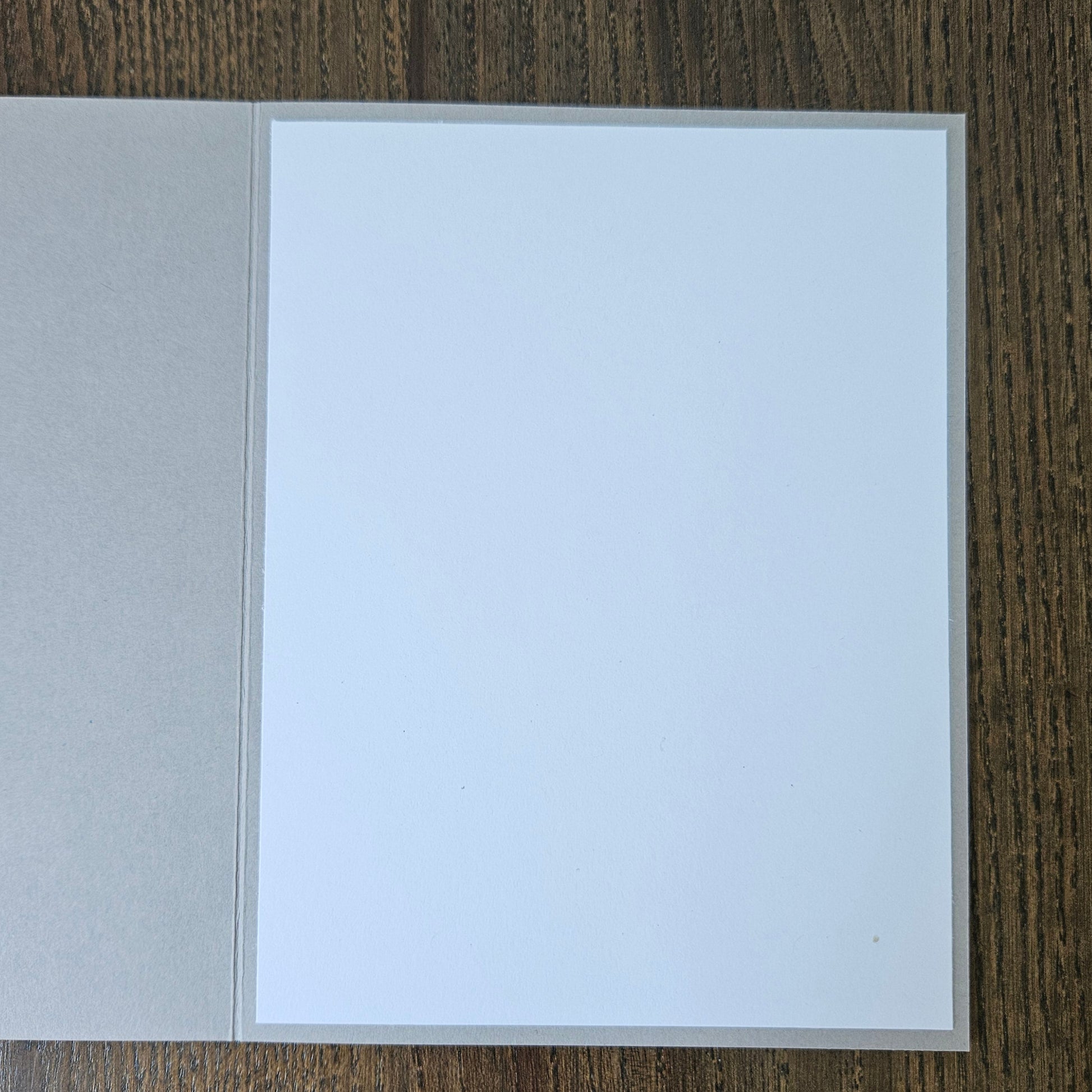 An open greeting card with a blank white center panel and a light gray textured left panel, photographed from above on a dark wood-grain surface.