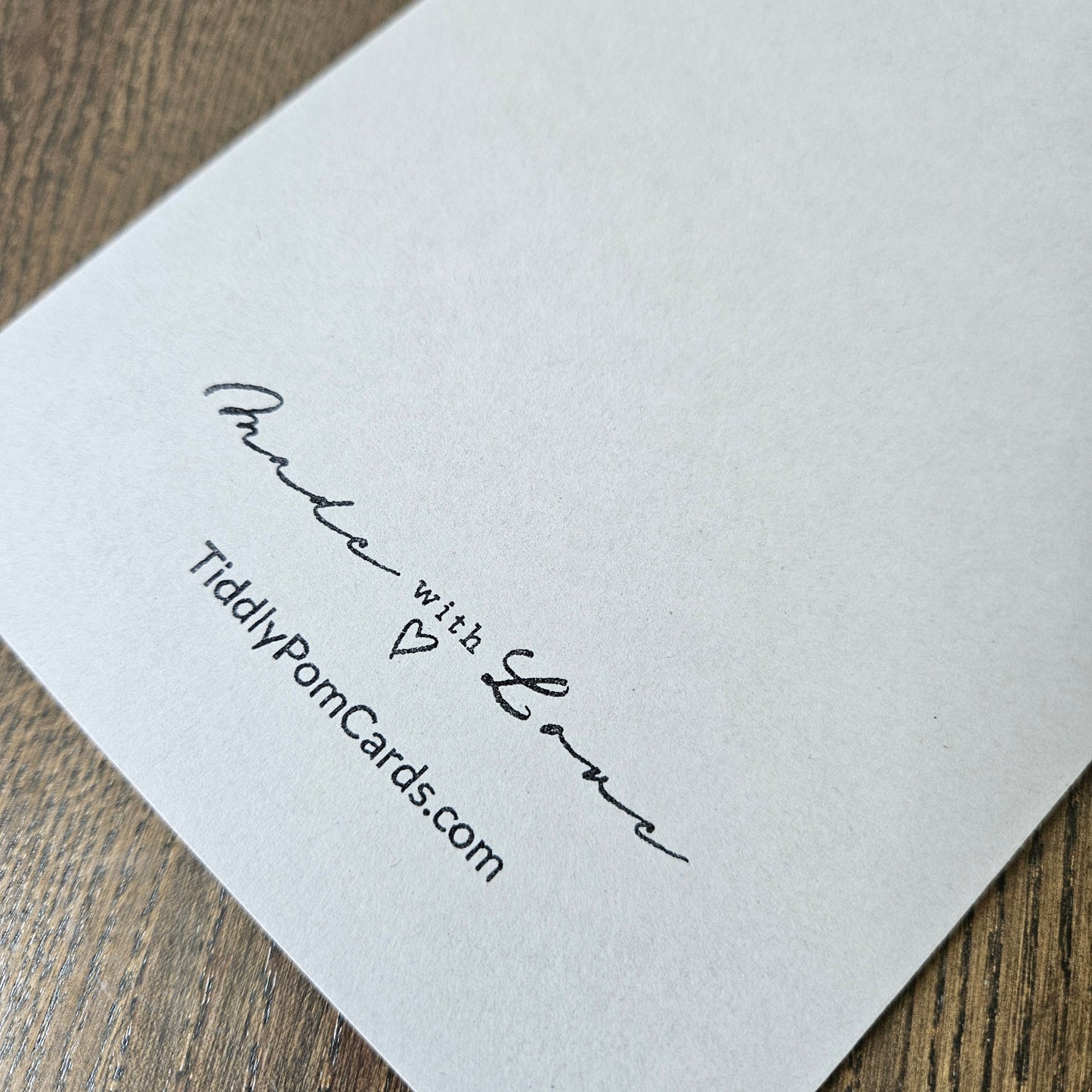 A close-up of a white textured card printed with the words 'Pude with Love' in black script, a small heart, and the website 'TiddlyPornCards.com', placed on a wooden surface.