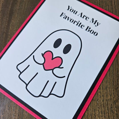 A handmade greeting card with a white background, a black and red border, and a cute ghost illustration holding a pink heart. The card is placed on a dark wood surface, with the text 'You Are My Favorite Boo' written at the top.