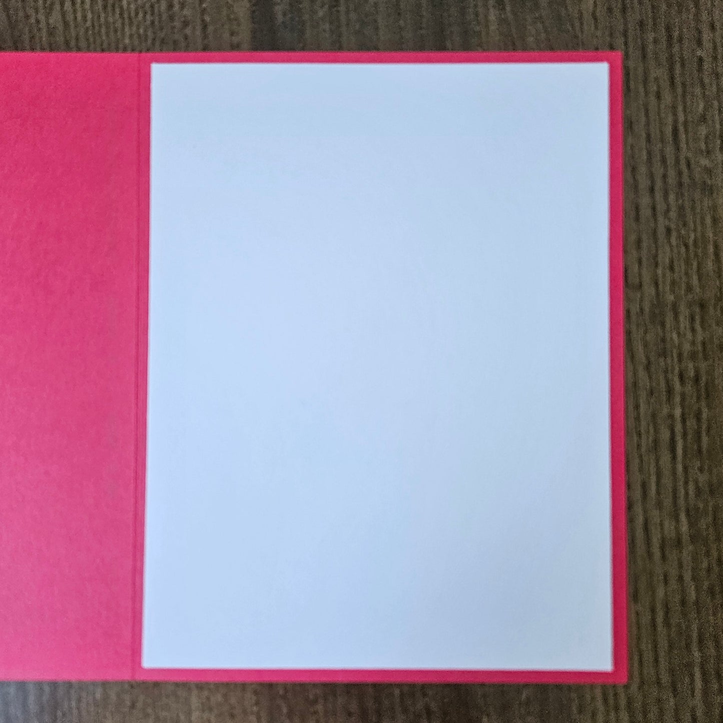 A blank white rectangular card or paper insert placed on a bright pink folder or book cover, photographed from above on a dark wood surface.