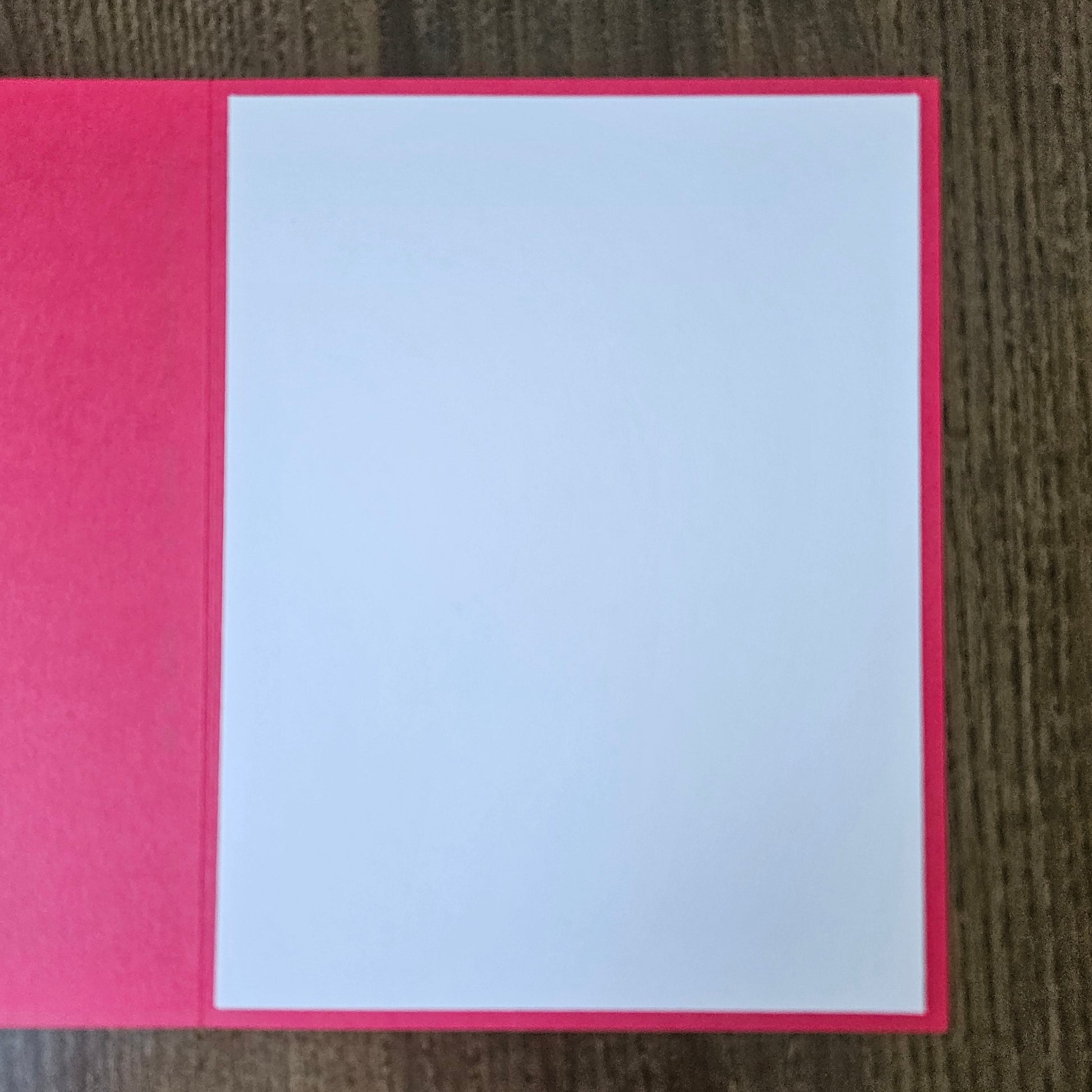 A blank white rectangular card or paper insert placed on a bright pink folder or book cover, photographed from above on a dark wood surface.