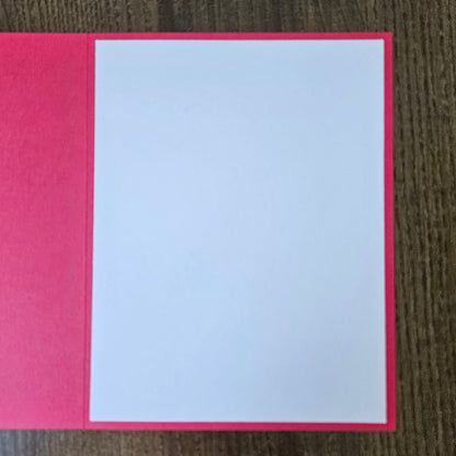 A blank white rectangular card or paper insert placed on a bright pink folder or book cover, photographed from above on a dark wood surface.
