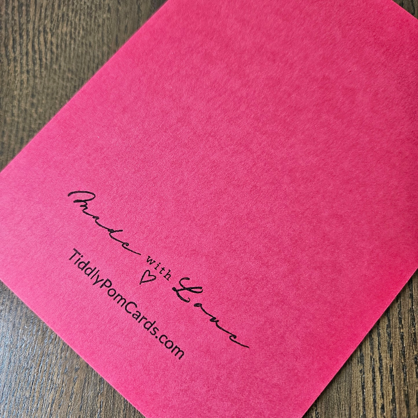 A bright pink textured pom pom card with black printed cursive text reading 'made with Love' and 'TidilyPomCards.com', shown on a dark wood surface.