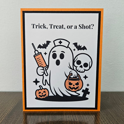 Front of a handmade A2 nurse Halloween card featuring a cute nurse ghost holding a syringe and pumpkin bucket framed by black and orange borders with text reading Trick Treat or a Shot.