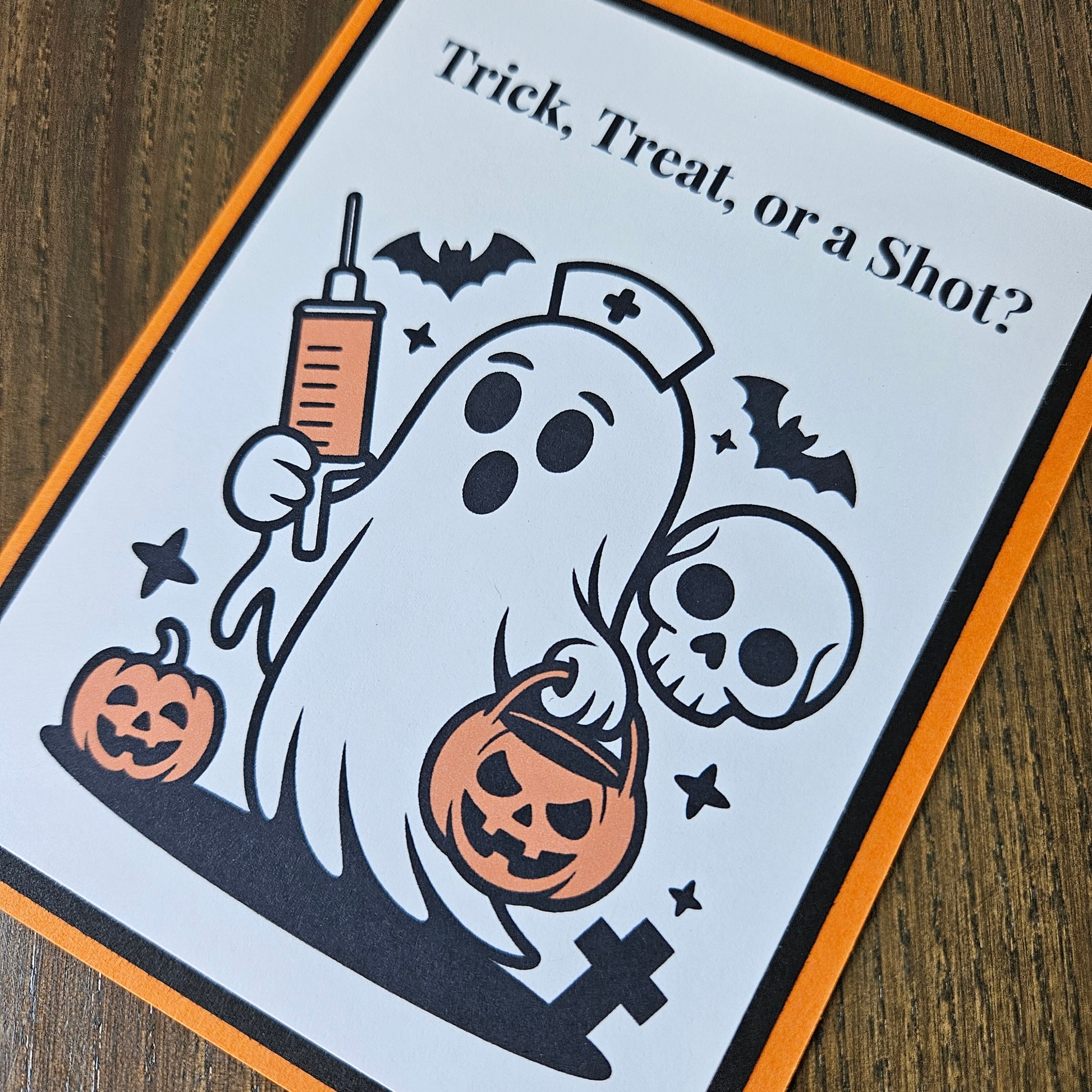 A Halloween-themed printed card or sign with the text 'Trick, Treat, or a Shot?' and a cartoon ghost holding a syringe and pumpkin, shown on a wooden surface.