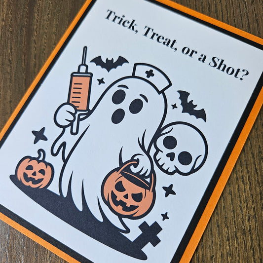 A Halloween-themed printed card or sign with the text 'Trick, Treat, or a Shot?' and a cartoon ghost holding a syringe and pumpkin, shown on a wooden surface.