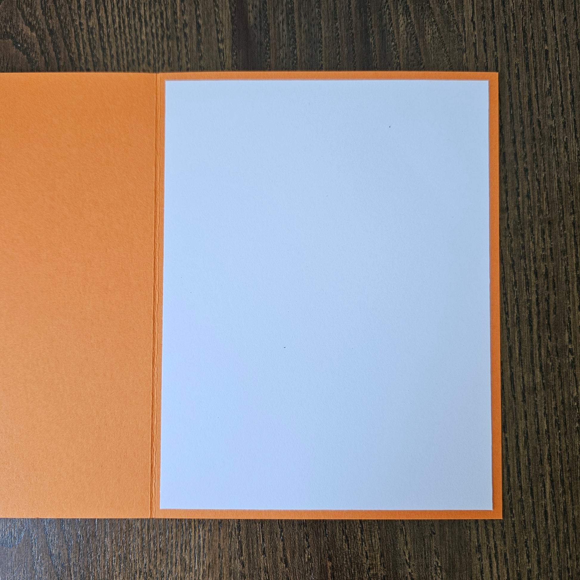 An open orange greeting card with a blank white center panel, displayed flat on a dark wood-grained surface.