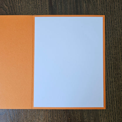 An open orange greeting card with a blank white center panel, displayed flat on a dark wood-grained surface.