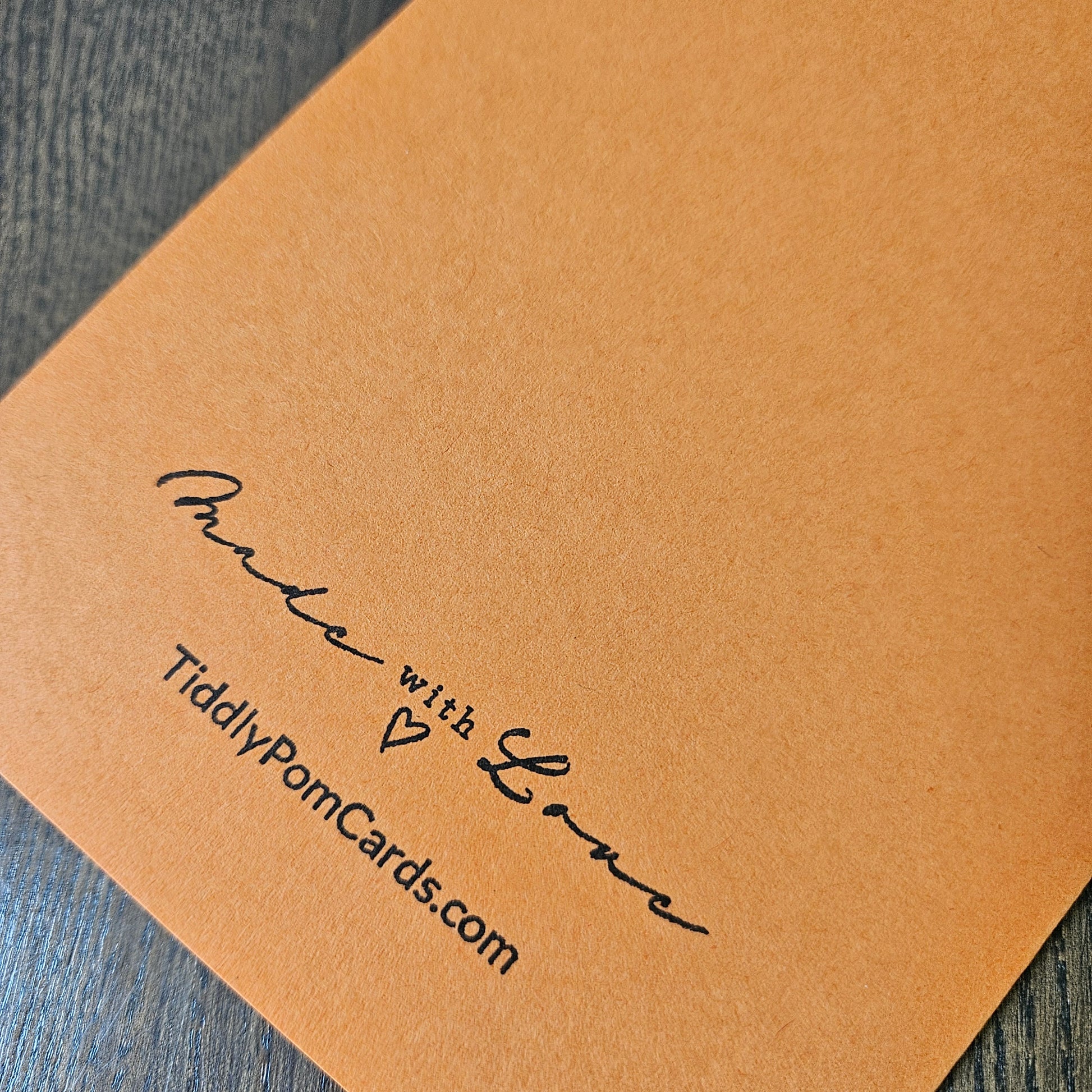 A close-up of a textured tan paper card printed with the black script 'Made with Love' and the website 'TiddlyPornCards.com', resting on a dark wood surface.
