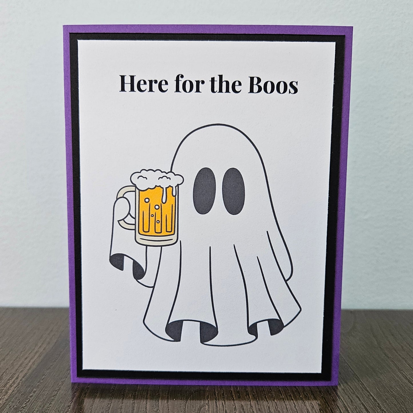 Front of a handmade A2 funny Halloween card featuring a ghost holding a beer mug framed by black and purple borders with text reading Here for the Boos.