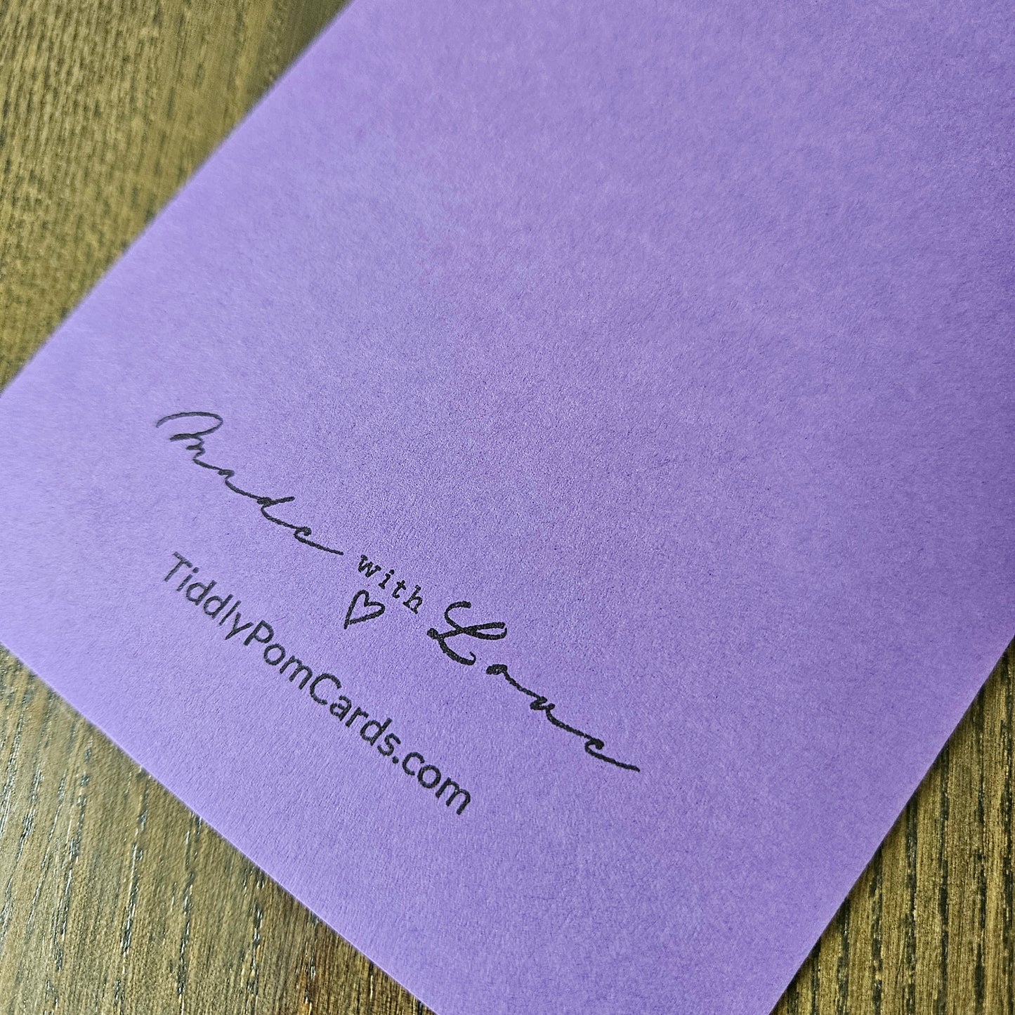 A close-up of a lavender purple textured card printed with 'Make with Love' and 'TidilyPomCards.com' in black script, resting on a wooden surface.