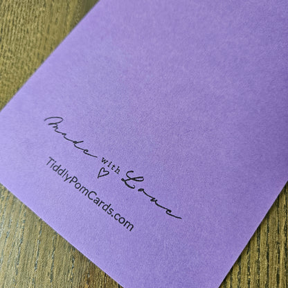 A close-up of a lavender purple textured card printed with 'Make with Love' and 'TidilyPomCards.com' in black script, resting on a wooden surface.