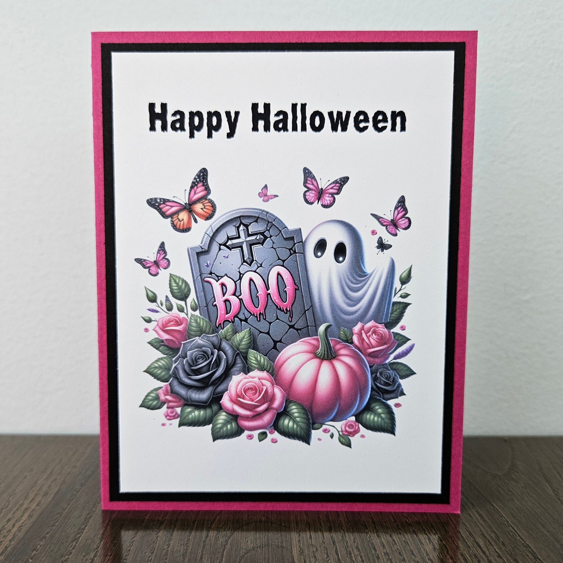 A handmade Halloween greeting card with a pink and black border, featuring a white background with the text 'Happy Halloween' and a colorful illustration of a tombstone with 'BOO', a white ghost, pink and gray pumpkins, roses, and butterflies, standing on a dark wooden surface against a light gray wall.