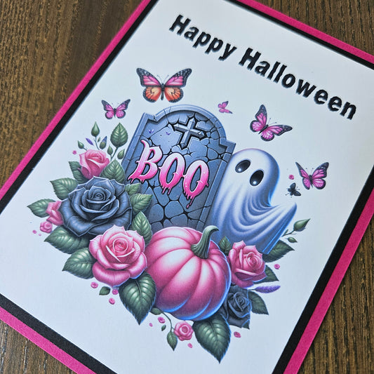 A Halloween greeting card with a white face, pink border, and black text reading 'Happy Halloween.' The card features a spooky illustration with a tombstone, ghost, pumpkins, roses, butterflies, and leaves on a wooden tabletop background.