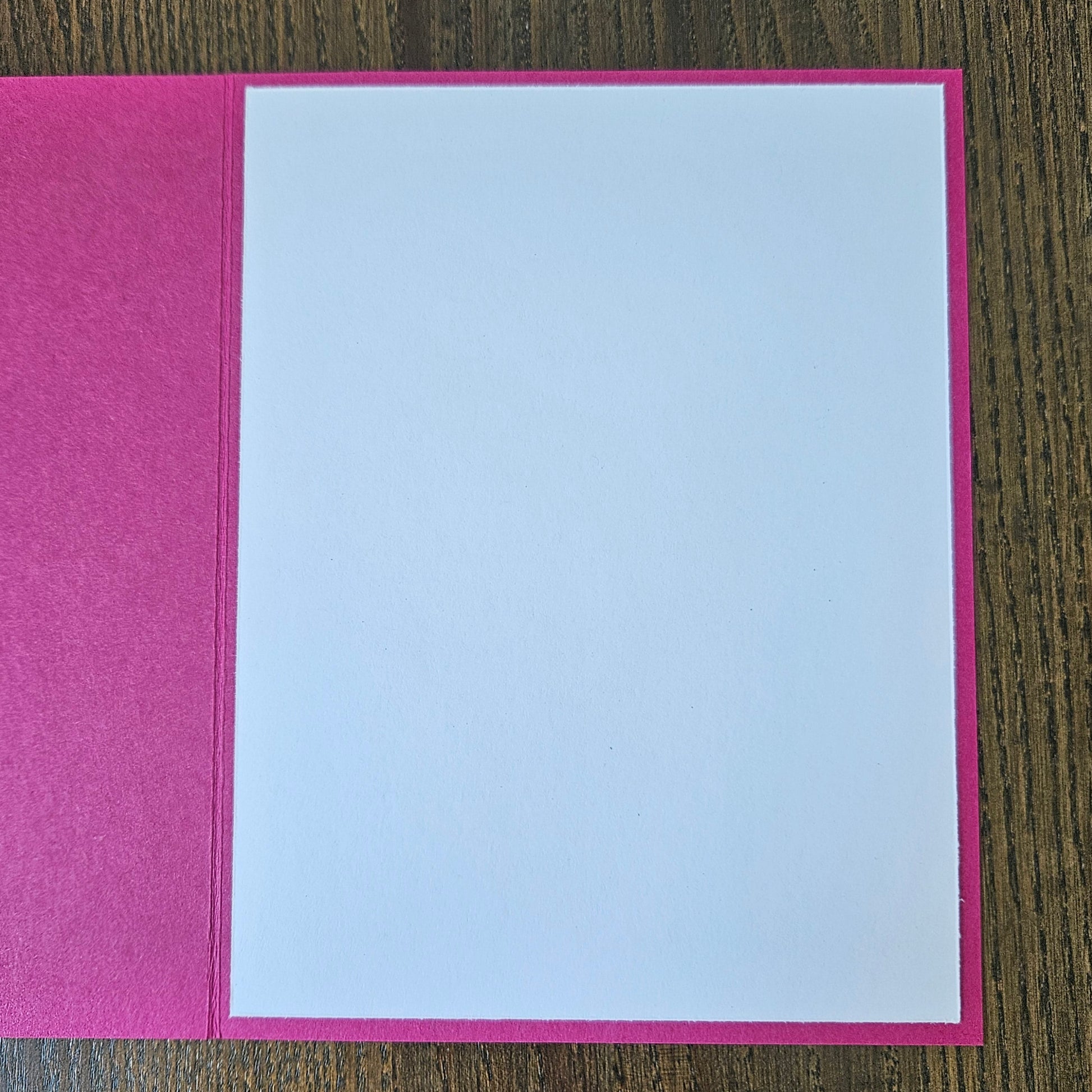 A greeting card with a textured hot pink cover and a blank white insert, shown open on a dark wood background.