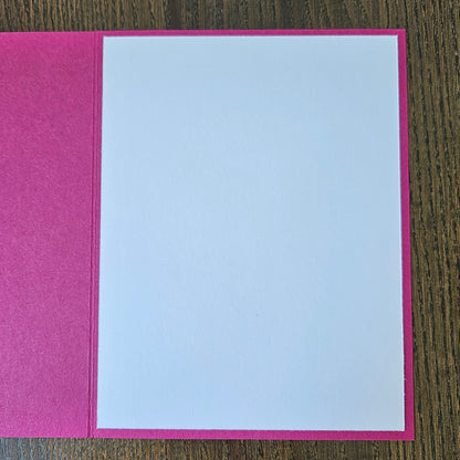 A greeting card with a textured hot pink cover and a blank white insert, shown open on a dark wood background.