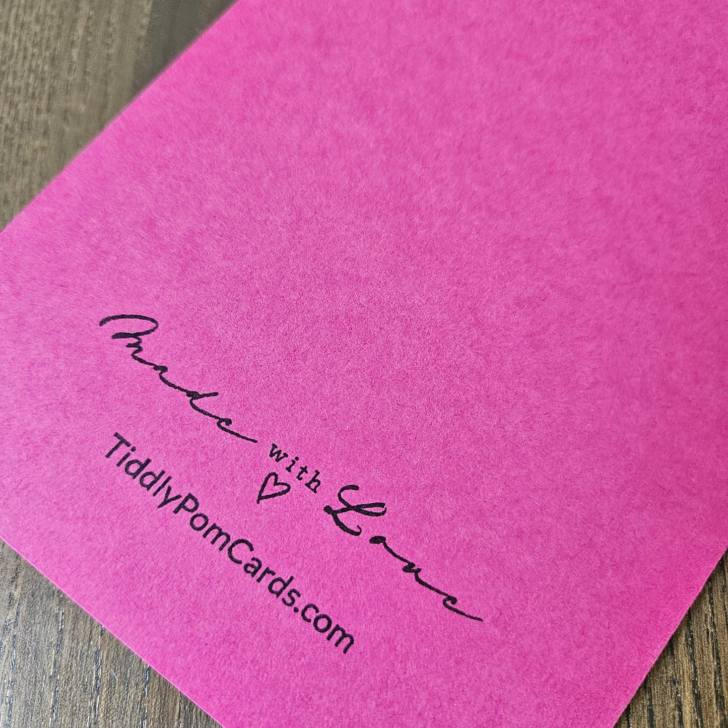 A close-up of a bright pink textured card or paper sheet with black printed text reading 'Made with Love' and 'TiddlyPornCards.com' on a wood surface.