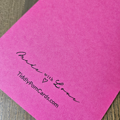 A close-up of a bright pink textured card or paper sheet with black printed text reading 'Made with Love' and 'TiddlyPornCards.com' on a wood surface.