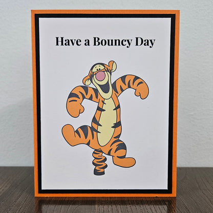 A square greeting card featuring Tigger from Winnie the Pooh standing on a spring with the text 'Have a Bouncy Day' above him. The card has an orange border, a black inner border, and a white center, displayed standing on a dark wooden surface against a light gray wall.