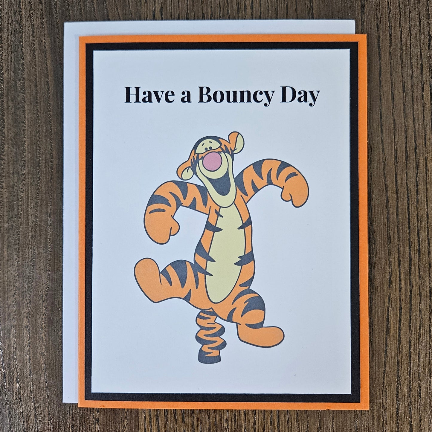 A square greeting card featuring the text 'Have a Bouncy Day' above a cartoon tiger character bouncing like a spring, styled with an orange and black border on a white card, placed on a dark wood background.