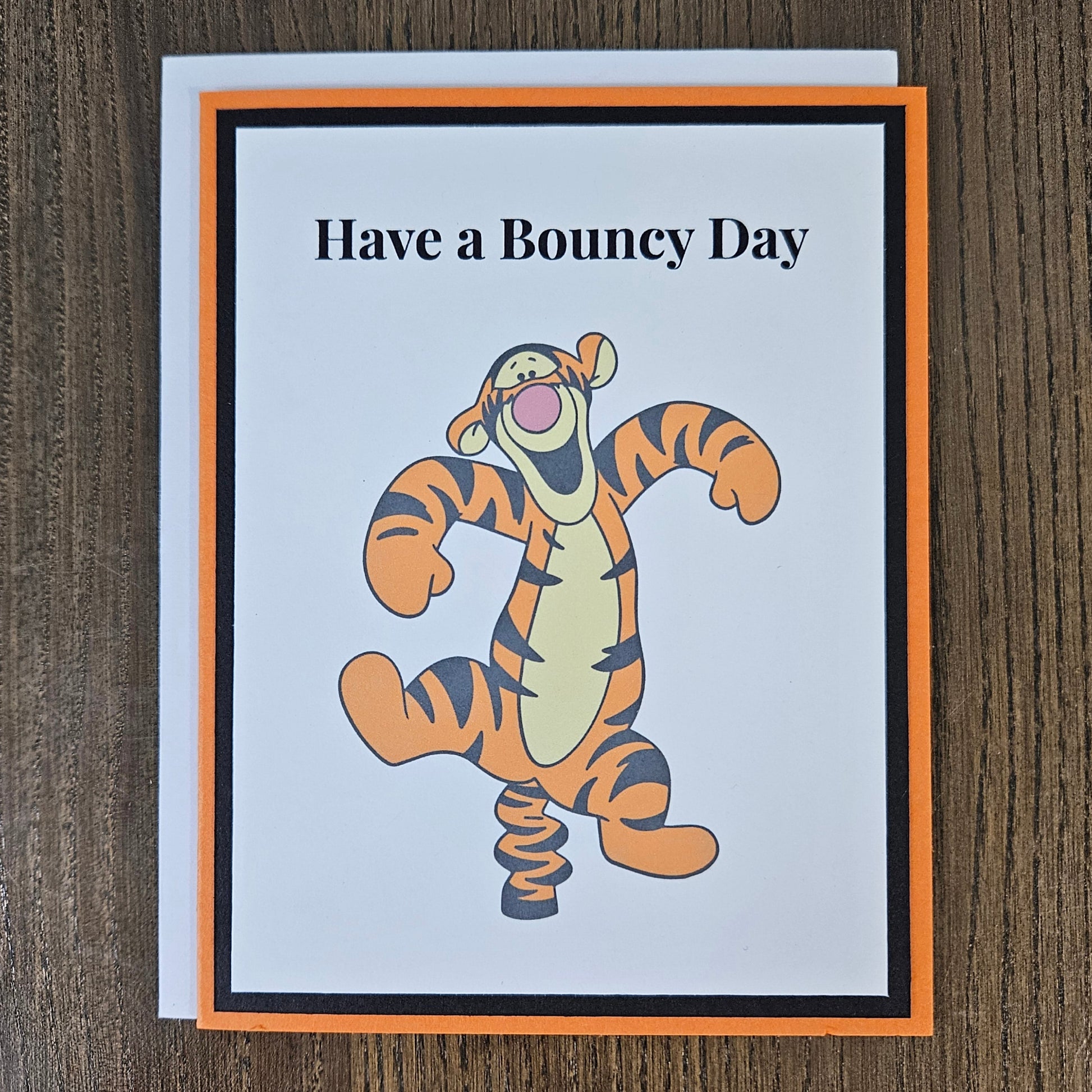 A square greeting card featuring the text 'Have a Bouncy Day' above a cartoon tiger character bouncing like a spring, styled with an orange and black border on a white card, placed on a dark wood background.