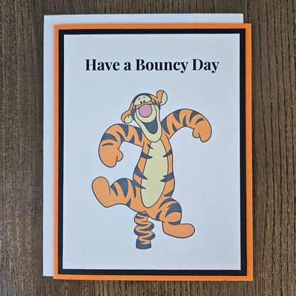 A square greeting card featuring the text 'Have a Bouncy Day' above a cartoon tiger character bouncing like a spring, styled with an orange and black border on a white card, placed on a dark wood background.