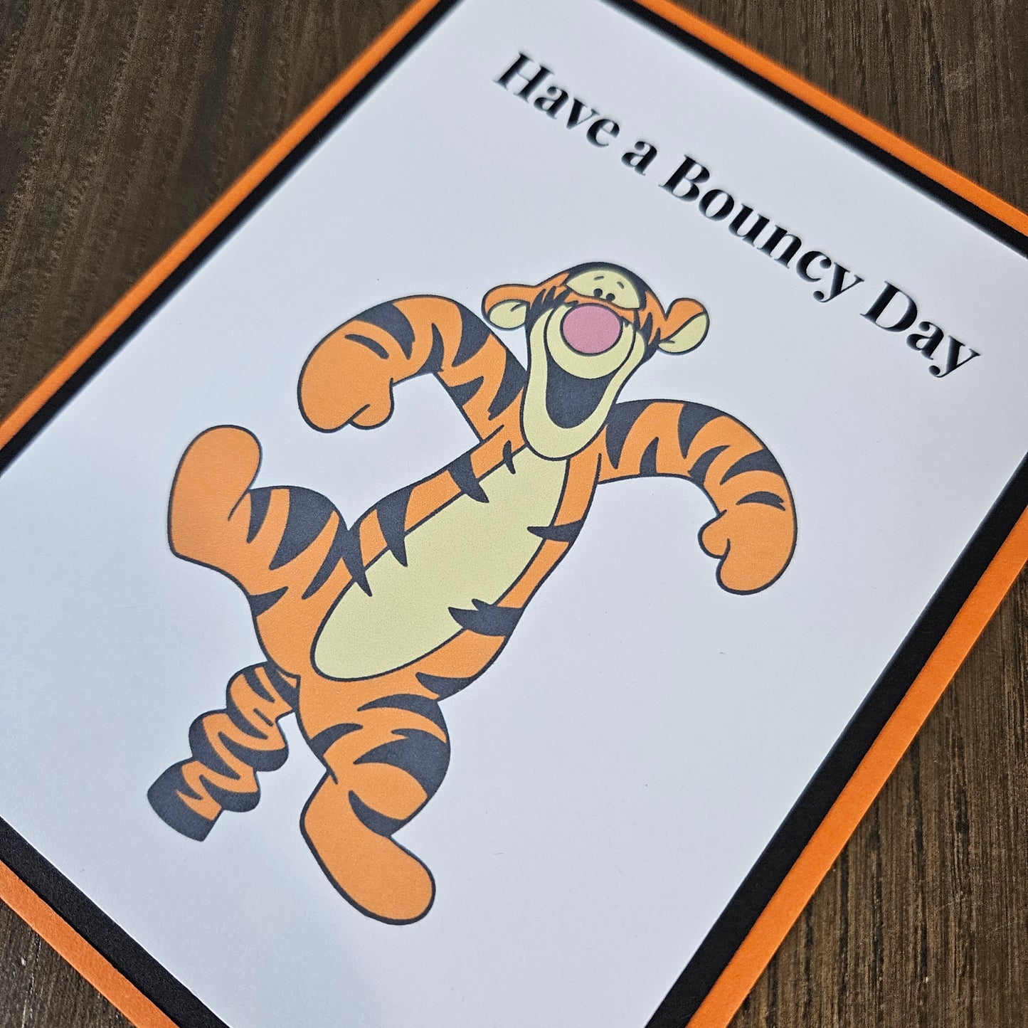 A square printed card or sign featuring Tigger from Winnie the Pooh holding a bouncing ball, with the text 'Have a Bouncy Day' at the top, placed on a dark wood surface.
