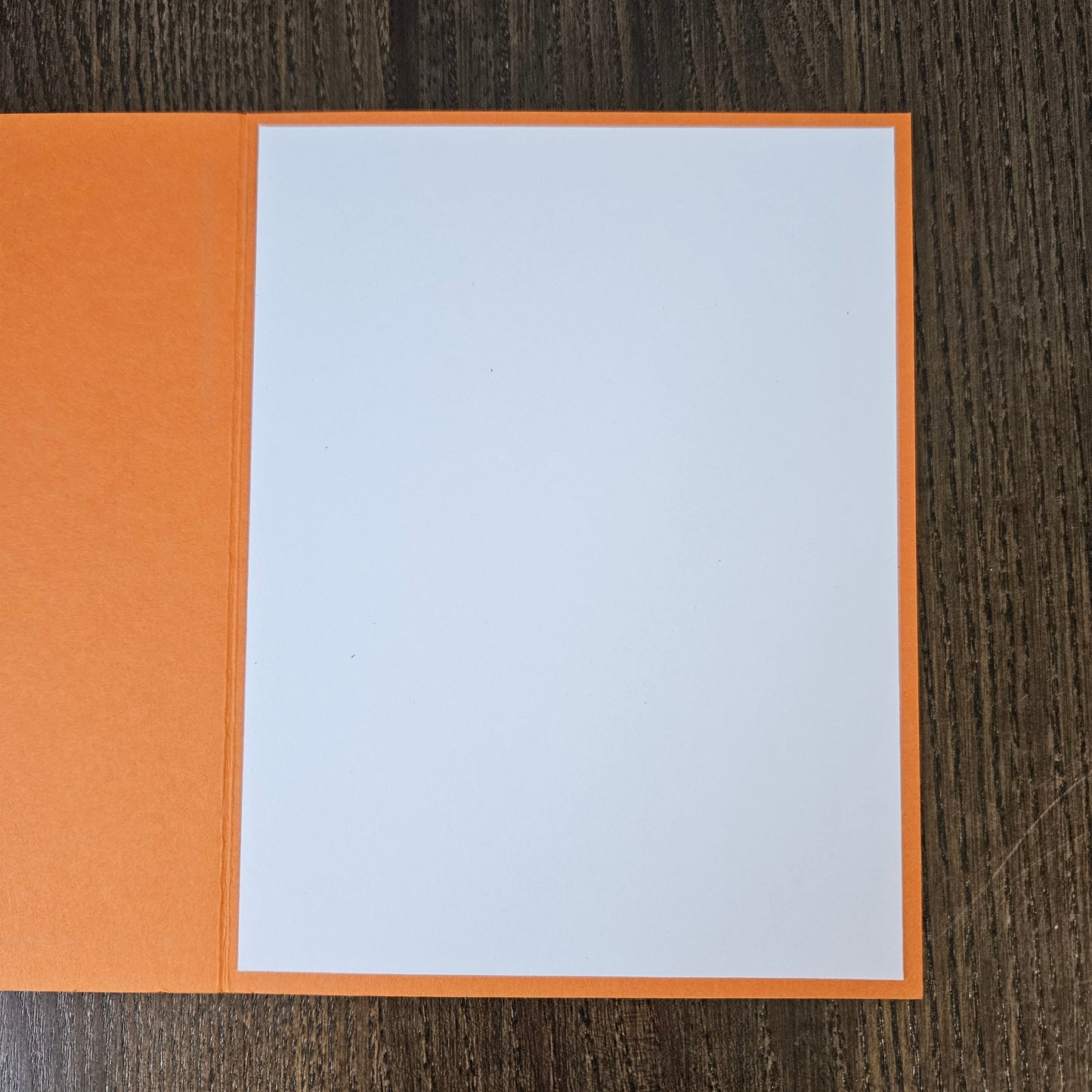 An open orange greeting card or notebook with a blank white page, photographed from above on a dark wood-grain surface.
